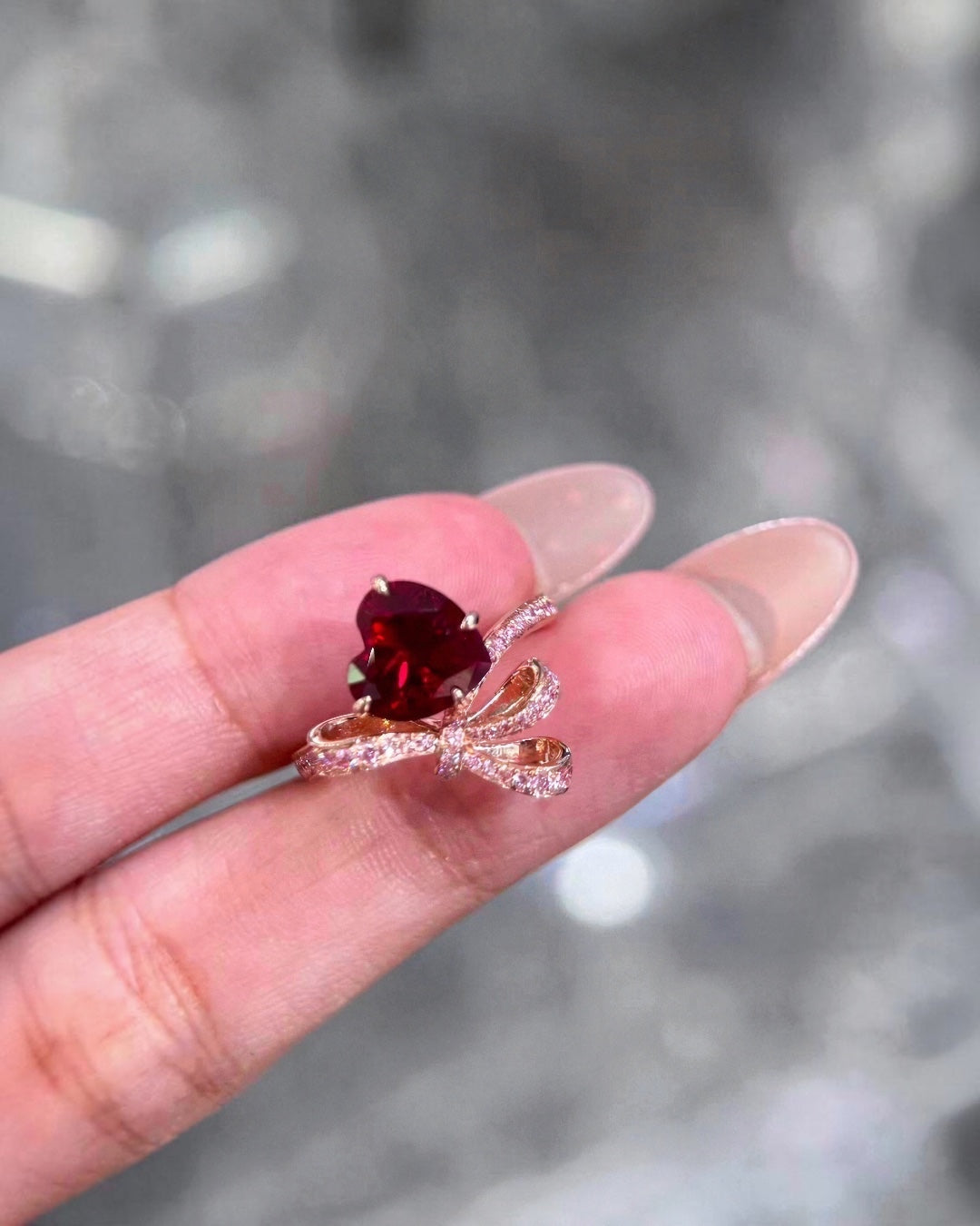 BoxUnboxed custom heart-shaped ruby ring with pink diamond accents in a gold bow setting worn on a finger