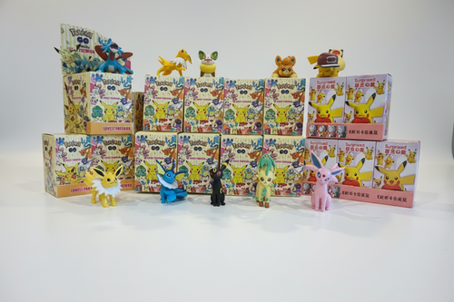 A collection of Pokémon TCG blind box sets and small character figures arranged on a white studio background. Multiple illustrated boxes with Pokémon artwork are stacked in rows, with several small toy figures placed in front and on top of the boxes.